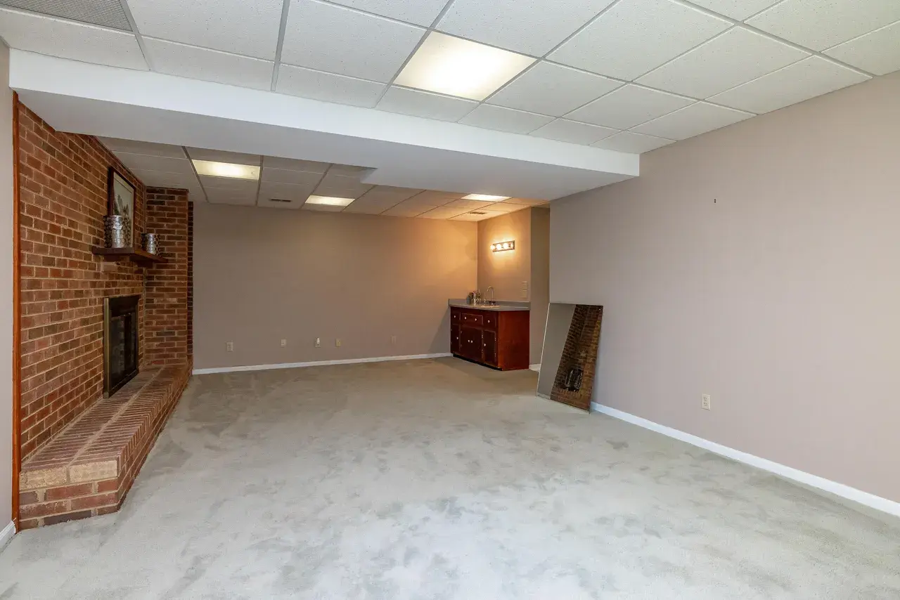 Basement before: empty room with carpet, a drop ceiling, and an exposed brick chimney breast.
