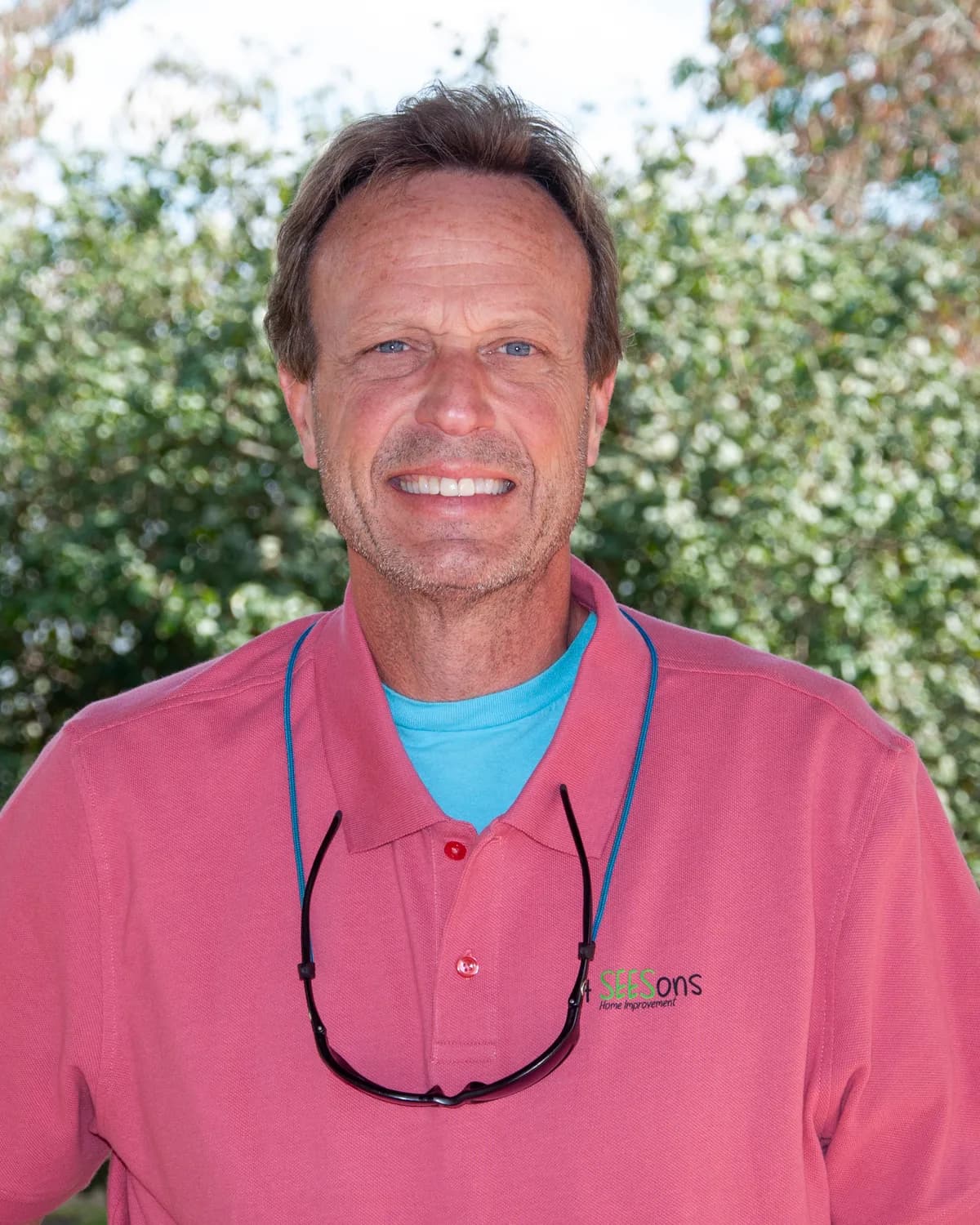 John Mark Stuart outdoors in front of green trees, smiling, wearing a 4SEESons polo shirt.